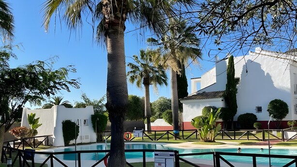 A swimming pool surrounded by a fence, palm trees, and a white building with a red trim.