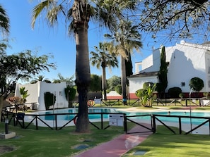 Piscina cercada por uma grade, palmeiras e um edifício branco com detalhes vermelhos.