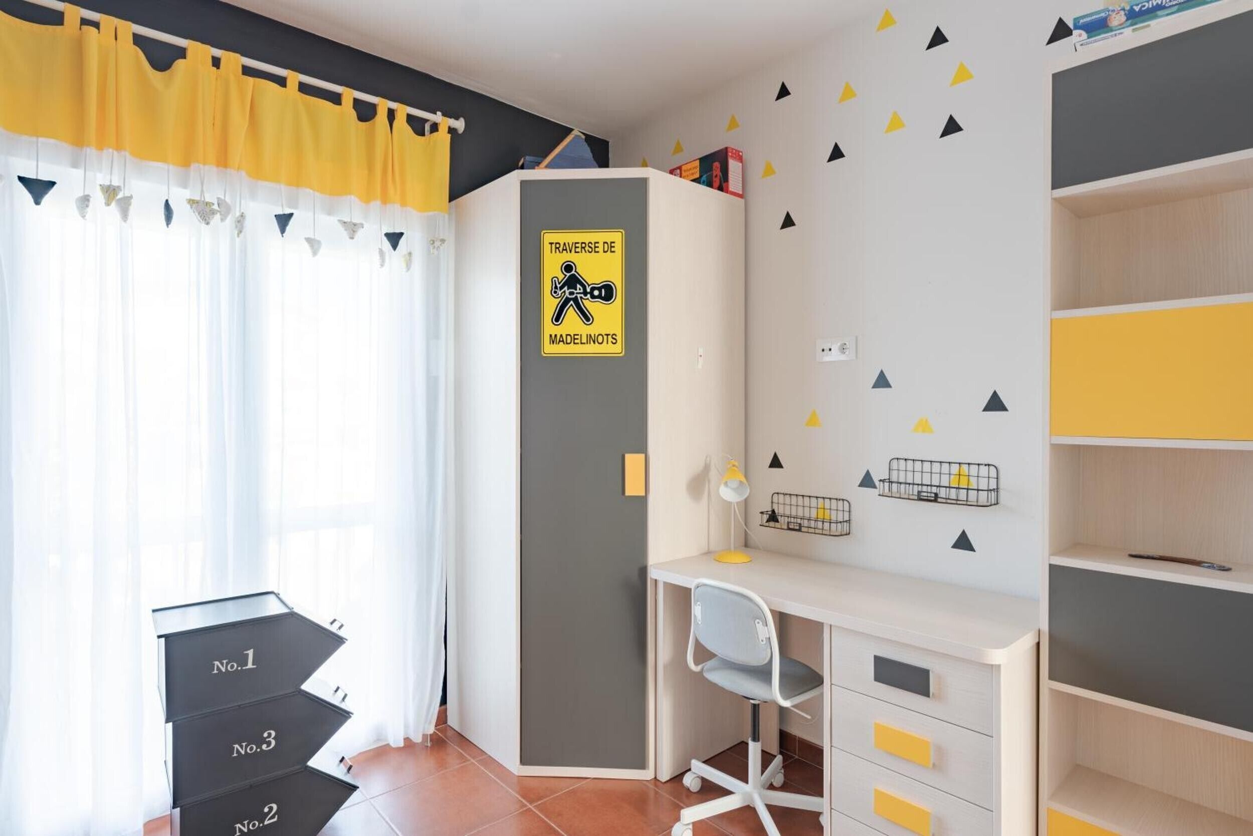 A room with a desk, chair, and storage units. There is a sign on the wall and a window with curtains.