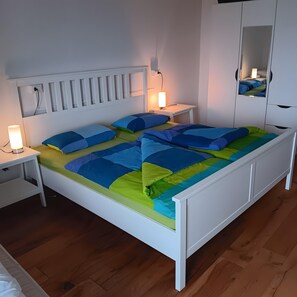 Un lit blanc avec une literie bleue et verte, une table de chevet avec une lampe et un miroir accroché au mur.