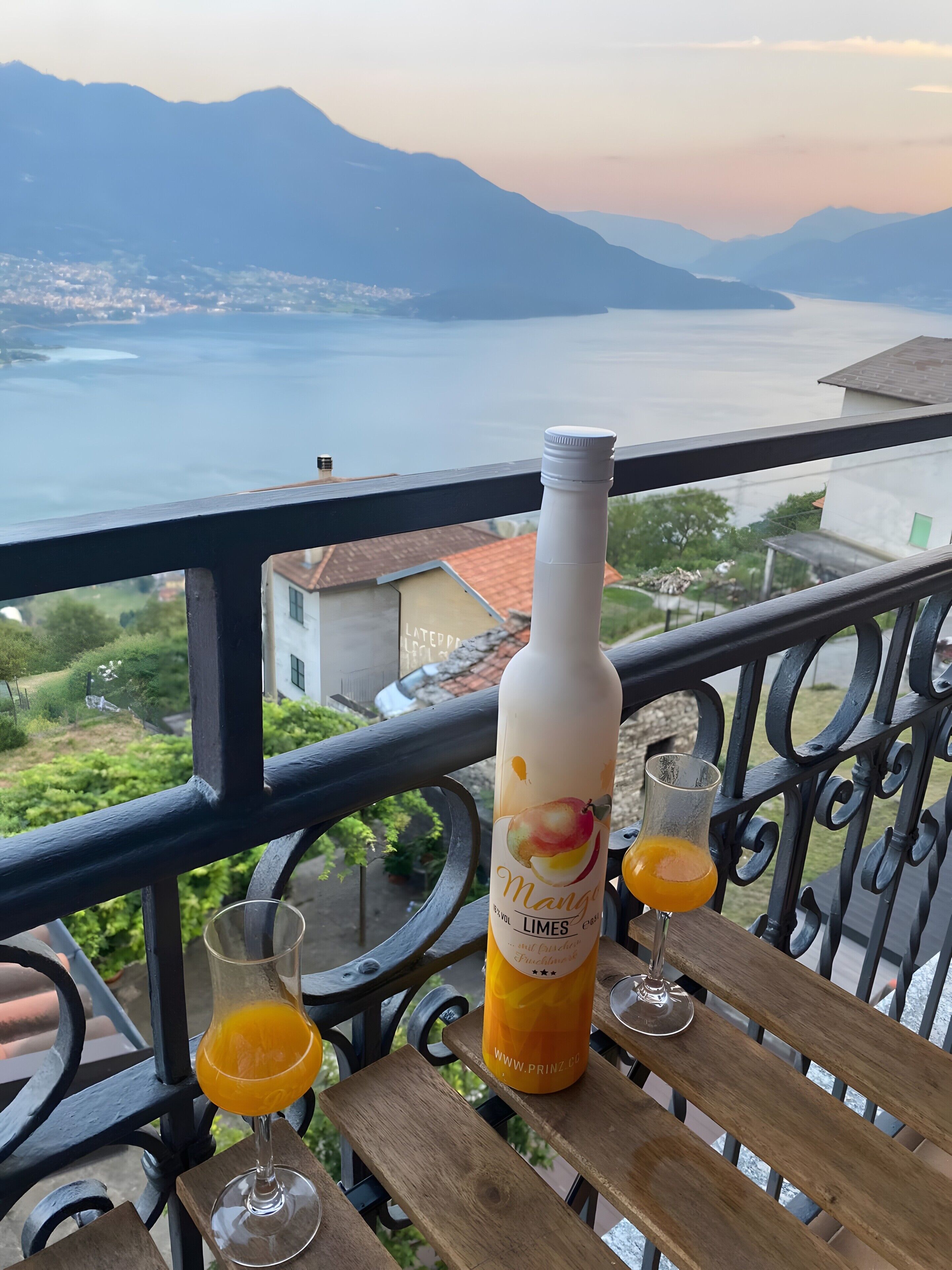 A bottle of mango limes on a balcony with two glasses of orange juice, overlooking a scenic view of mountains and a body of water.