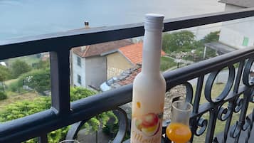 Een fles mangolimoen op een balkon, met twee glazen sinaasappelsap, met uitzicht op een schilderachtig berglandschap en een wateroppervlak.
