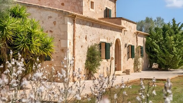 A stone building with arched windows and a central doorway, surrounded by greenery and flowering plants.
