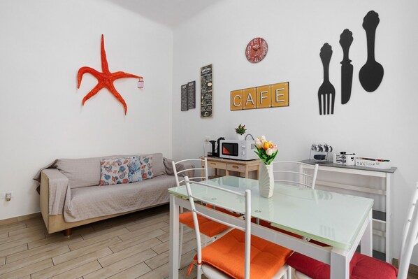 Un salon moderne avec une table à manger en verre, des chaises blanches, un canapé beige, une étoile de mer rouge en guise de décoration, une horloge murale et un panneau indiquant « CAFÉ ».