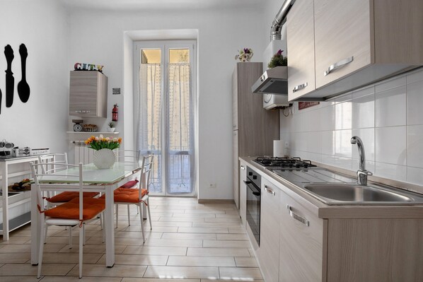 A modern kitchen with a gas stove, sink, and white cabinetry. There is a dining area with a glass table and orange chairs. - Apartment 'Ludy' with Balcony, Wi-Fi and Air Conditioning (La Spezia)