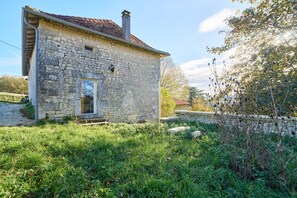 Une maison en pierre avec un toit en pente, un petit porche et une cour couverte d’herbe.