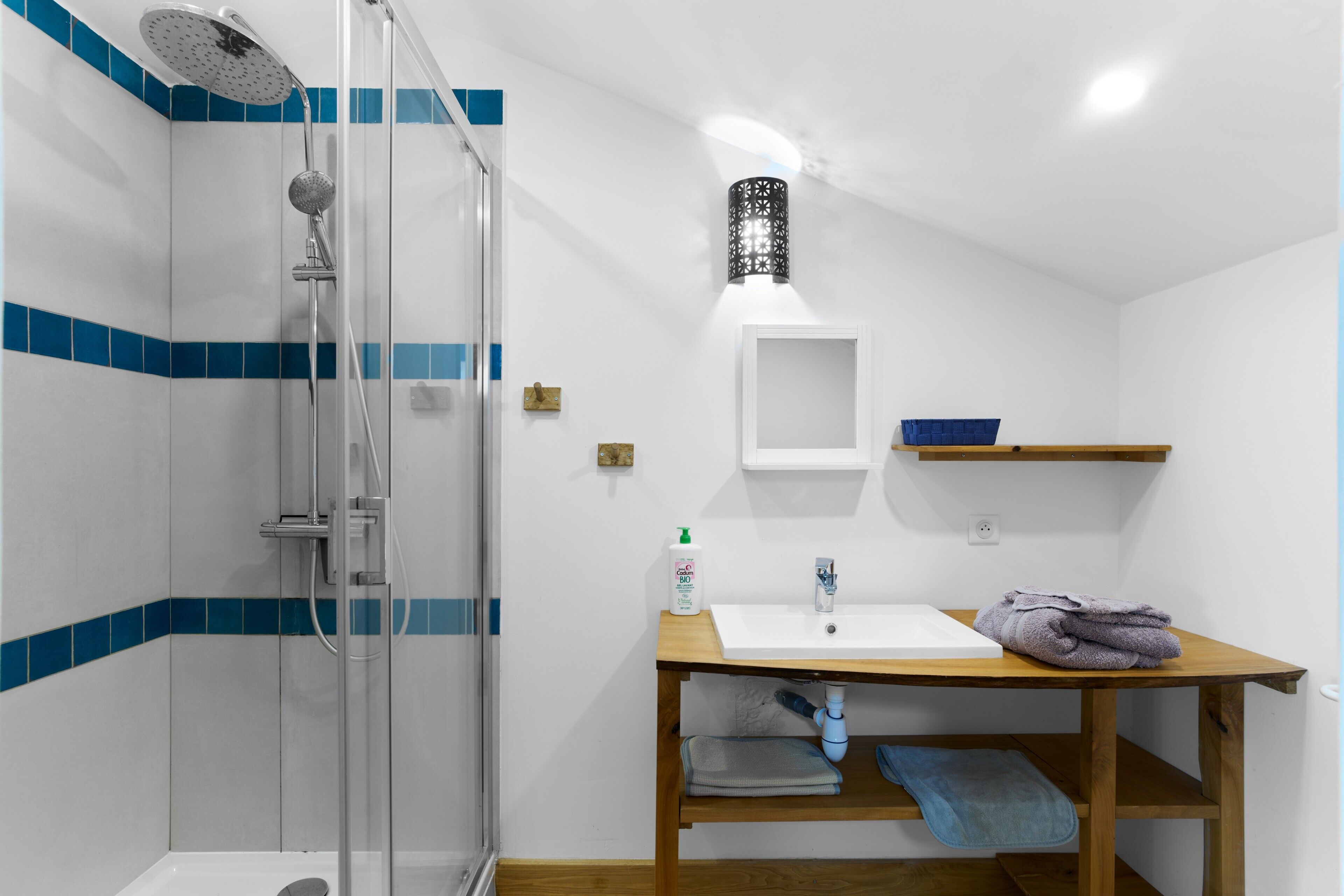 Une salle de bain avec une douche à parois de verre, un lavabo avec un miroir et une étagère en bois où sont posées des serviettes.