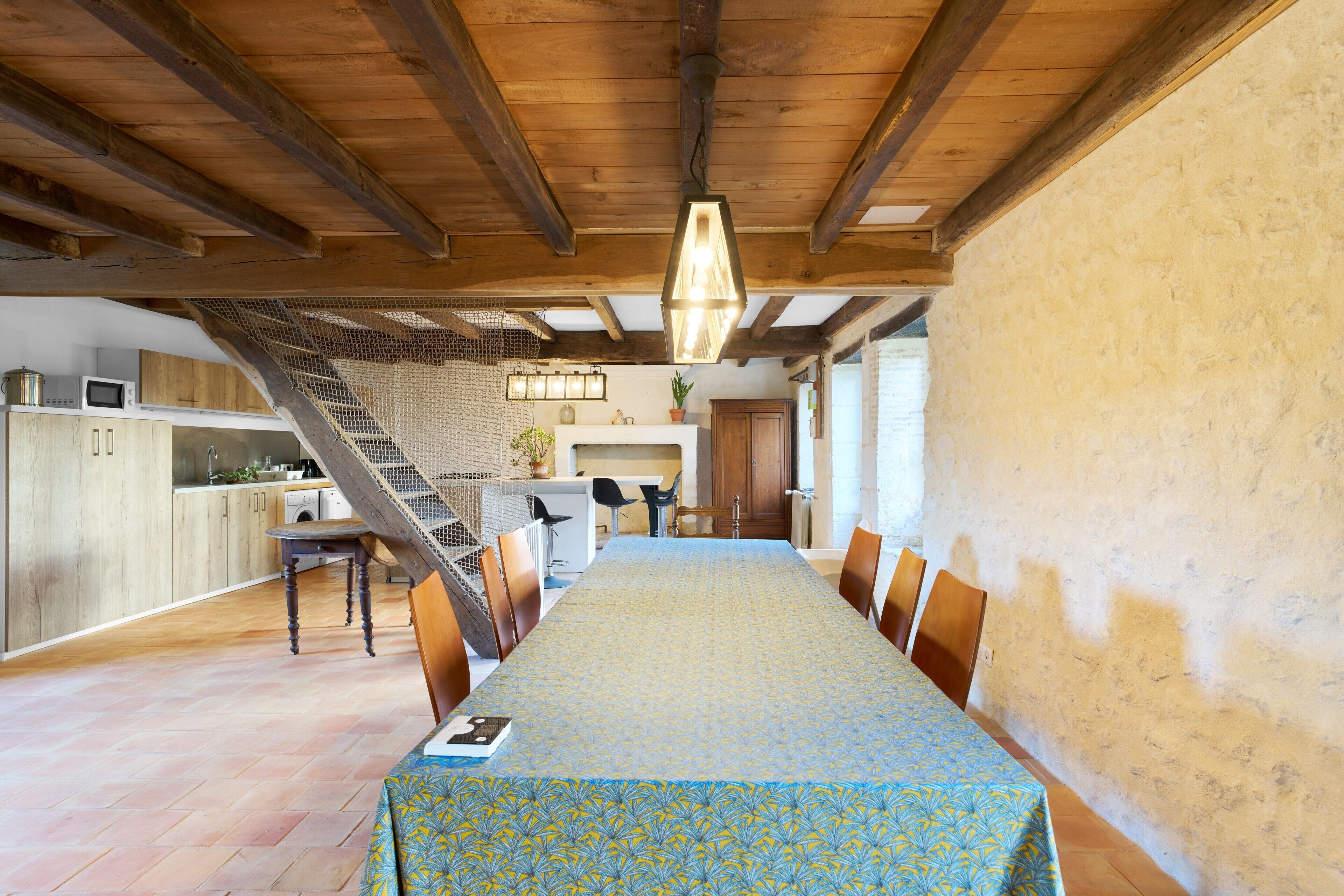 A dining area with a long table, chairs, a staircase, and a kitchen in the background.
