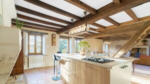 A kitchen with wooden beams, a central island, bar stools, and a view of the outdoors. - Traditional and Organic Gîte in the Tude Valley – Nature, Comfort, and Relaxation (Montboyer)