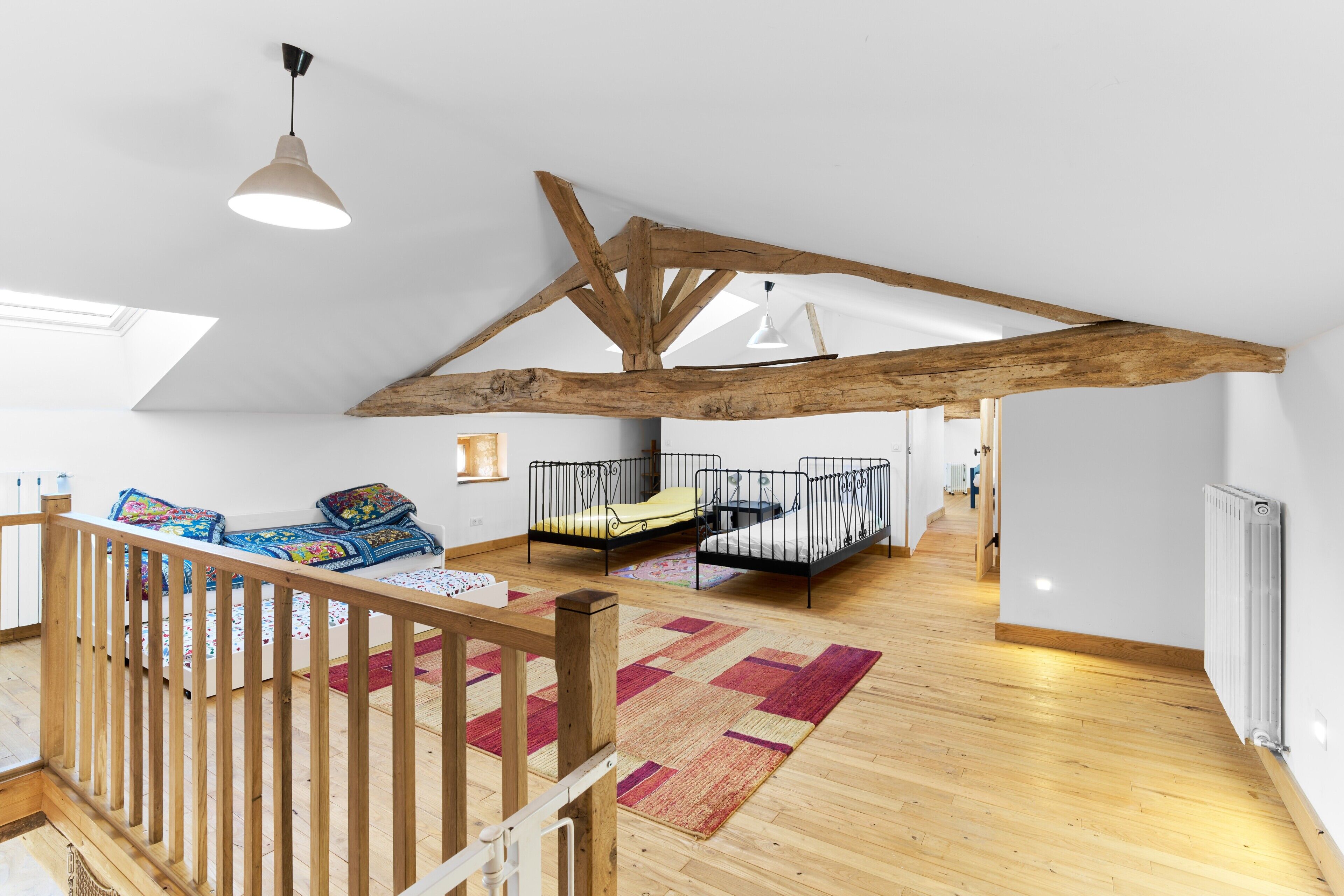 A spacious loft bedroom with a high ceiling, exposed wooden beams, a wooden staircase, and a large window.