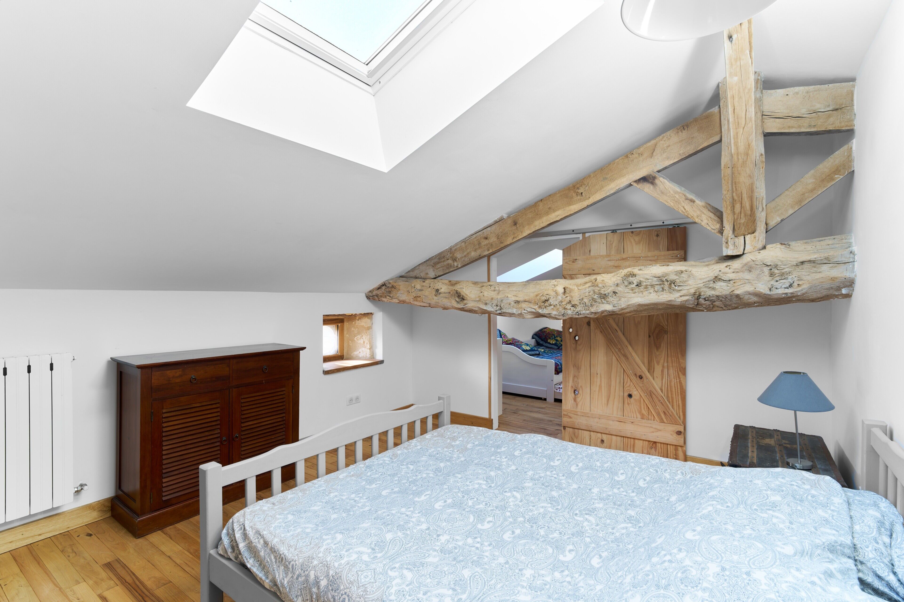 Une chambre avec un plafond en pente, des poutres en bois, un lit, une armoire, un radiateur et un puits de lumière.