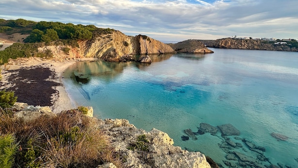 A coastal landscape with clear waters, rocky cliffs, and a distant shoreline with buildings.