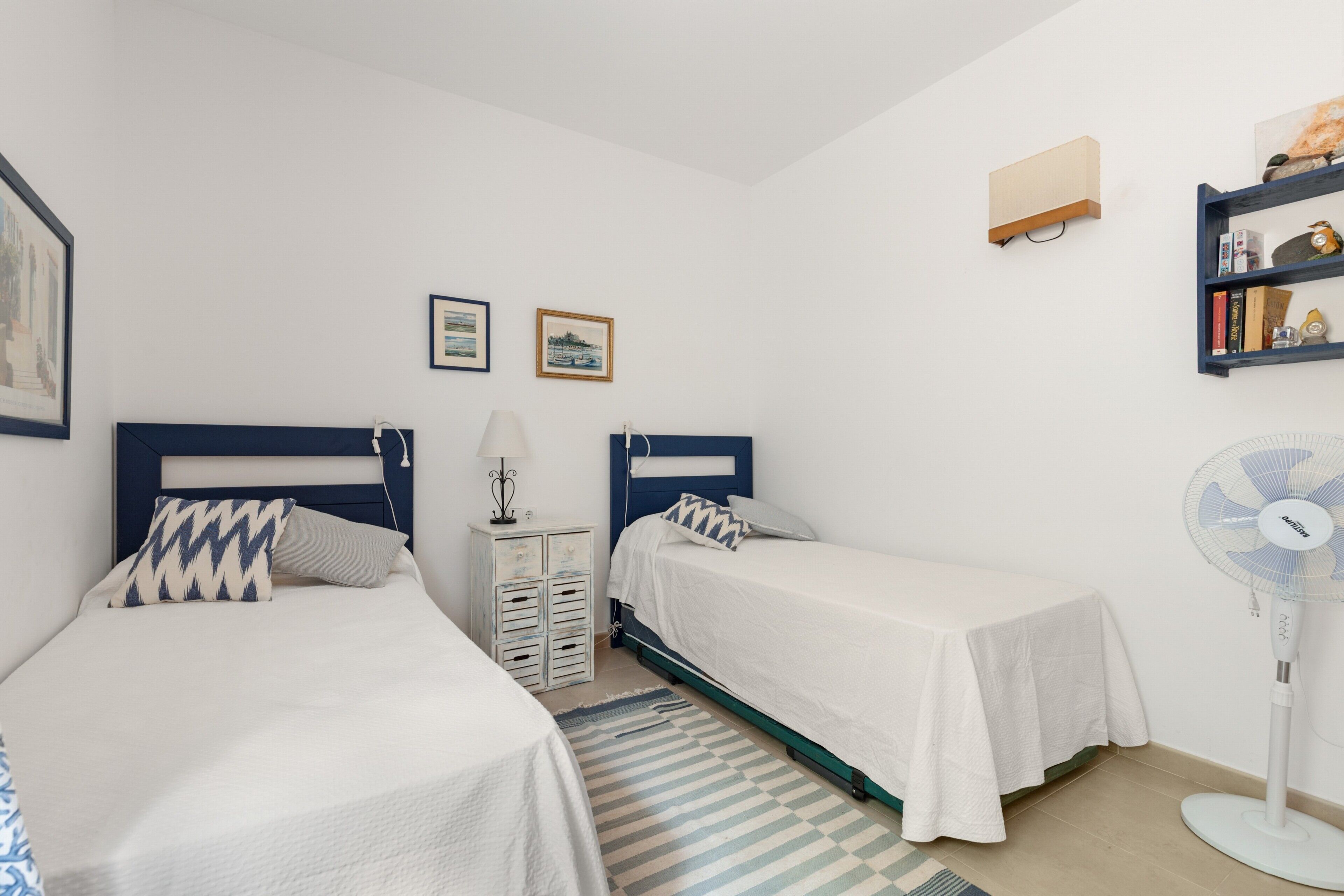 A bedroom with two beds, a bedside table, a lamp, a fan, and a bookshelf.
