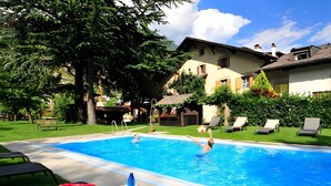 Una piscina con sillones de descanso, una mesa con frutas y bebidas, y una casa al fondo.