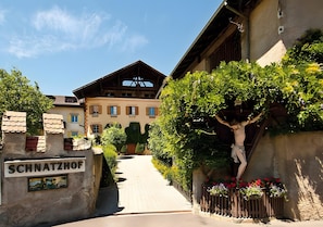 A building with a sign reading 'SCHNATZHOF,' a wooden structure with a crucifix, and a pathway leading to a building with a sloped roof.
