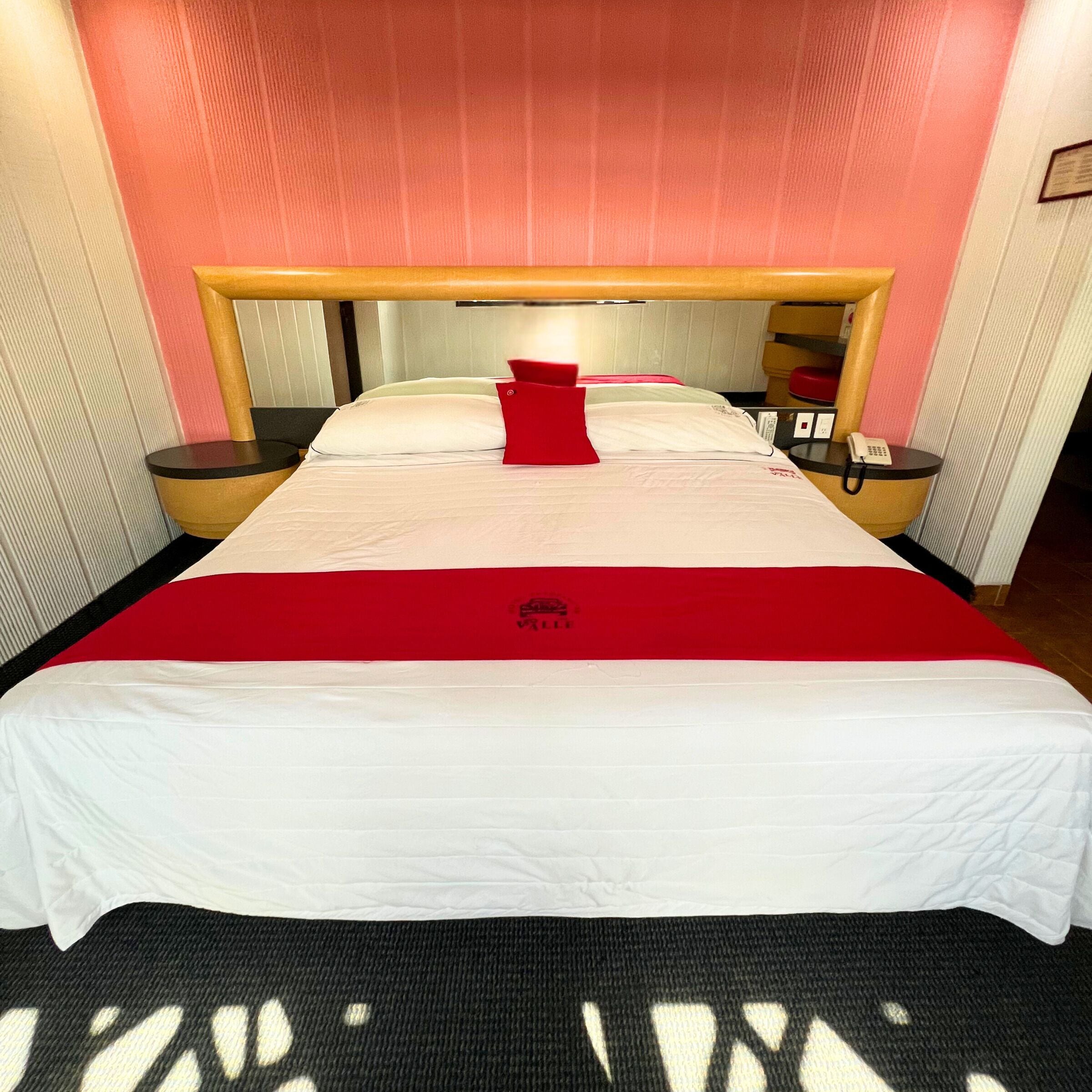 A hotel room with a large bed, a red throw pillow, and a bedside table with a phone.