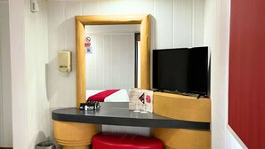 Una habitación de hotel pequeña con una cama, un televisor, una mesita y un taburete rojo.