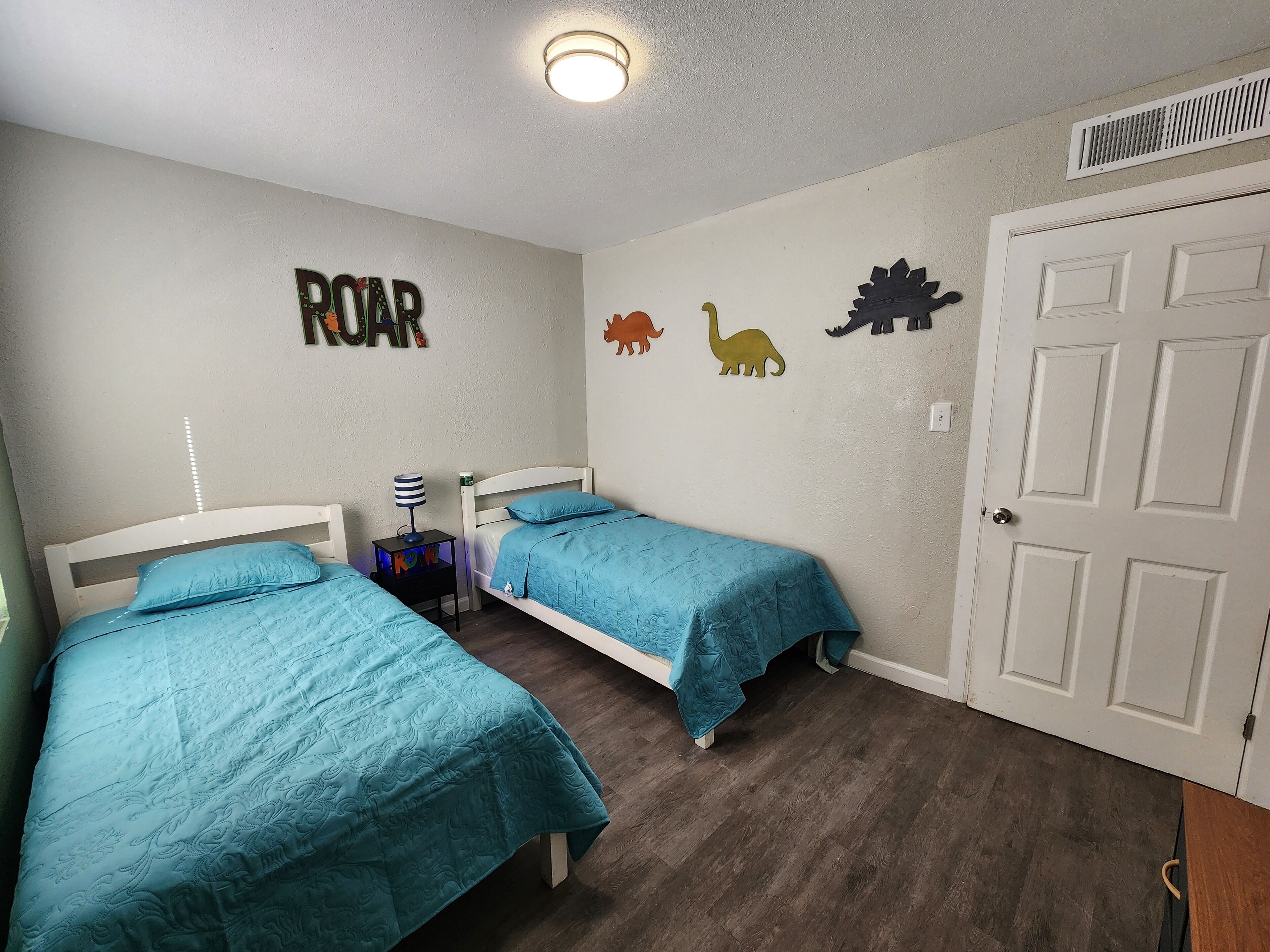 Une chambre avec deux lits, chacun recouvert d’une couverture bleue, une table de chevet avec une lampe et une décoration murale représentant des figurines de dinosaures.
