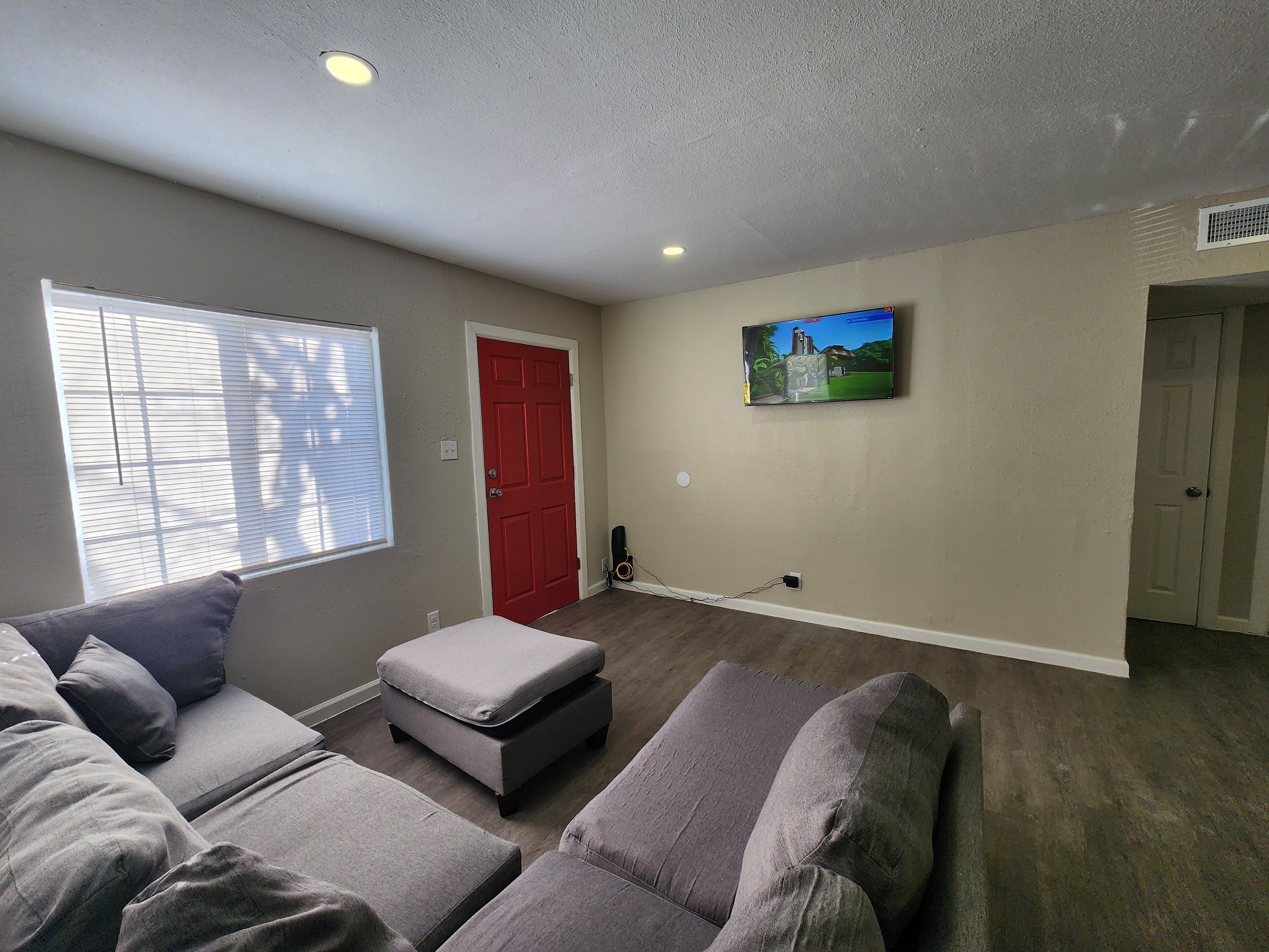 A living room with grey sofas, a red door, a wall-mounted TV, and a window with blinds.