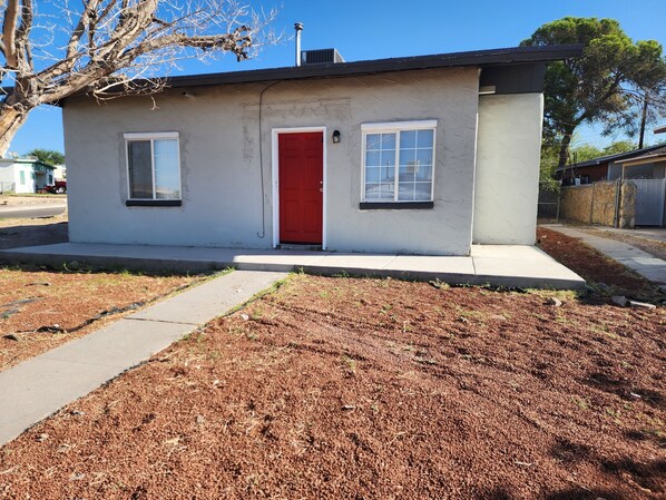 A small, single-story building with a red door and two windows, surrounded by a landscaped area with mulch and a sidewalk.