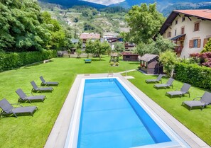 Une piscine rectangulaire entourée d’une pelouse, avec des chaises longues et une vue sur un paysage montagneux.