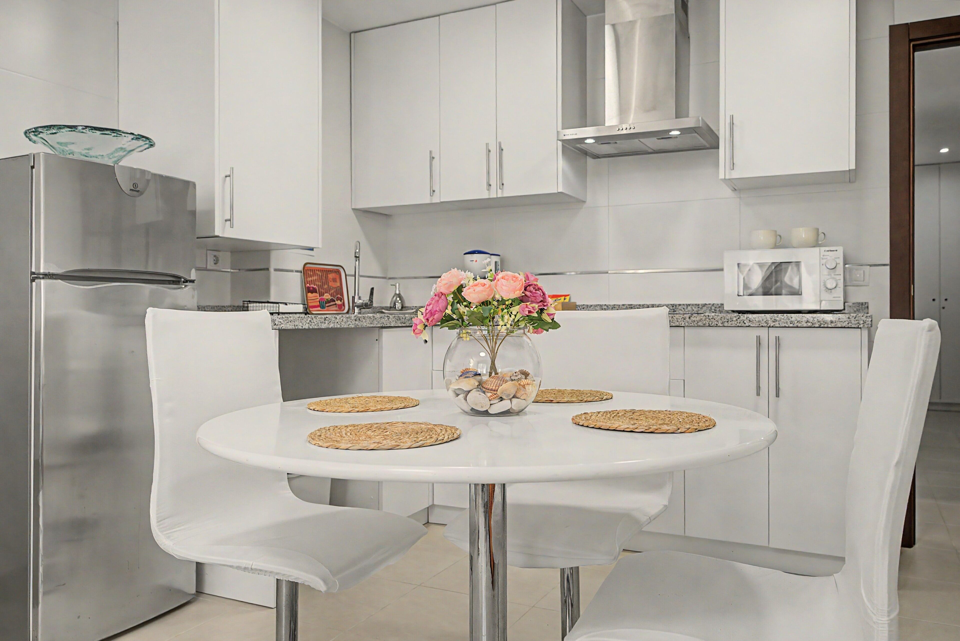 A modern kitchen with white cabinetry, a stainless steel refrigerator, a microwave, and a dining table with chairs.