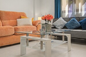A modern living room with a glass coffee table, orange sofa, and grey sofa with blue cushions.