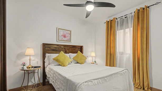 Quarto com cama, duas luminárias de cabeceira, ventilador de teto, janela com cortinas e um quadro na parede.
