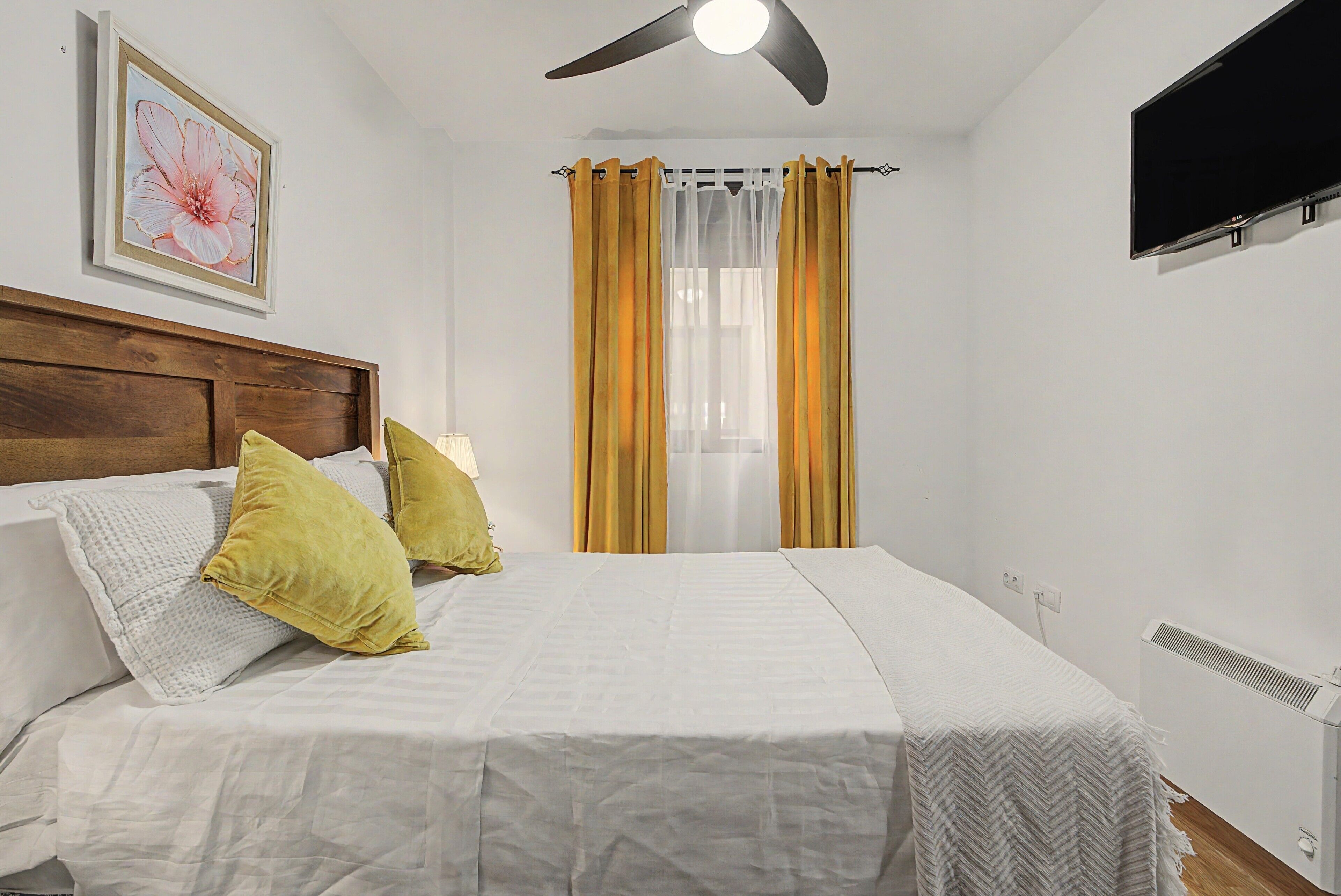 Una habitación de hotel con una cama, cabecera, televisor montado y un cuadro en la pared.