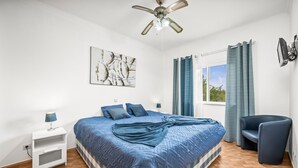 Ein Schlafzimmer mit einem Bett, einem Nachttisch, einem Sessel, einem Deckenventilator, einem Fenster mit Vorhängen und einem an der Wand montierten Fernseher.