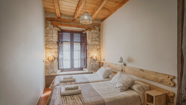 Une chambre avec deux lits, des murs en pierre, des poutres en bois, un luminaire suspendu et une fenêtre avec des volets.