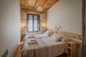 Un dormitorio con dos camas, paredes de piedra, vigas de madera, una lámpara colgante y una ventana con contraventanas.