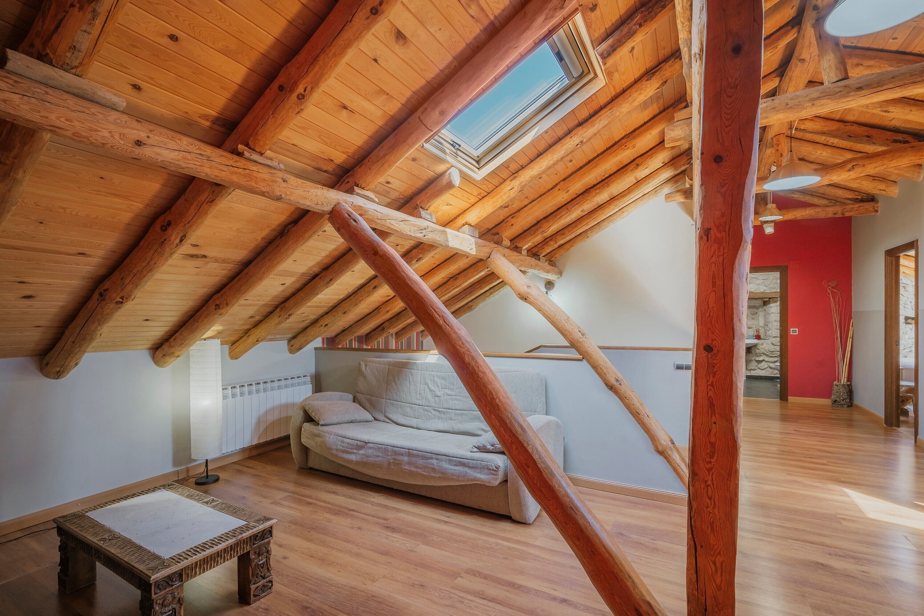 A loft bedroom with a sloped ceiling, wooden beams, a skylight, a sofa, and a small table.