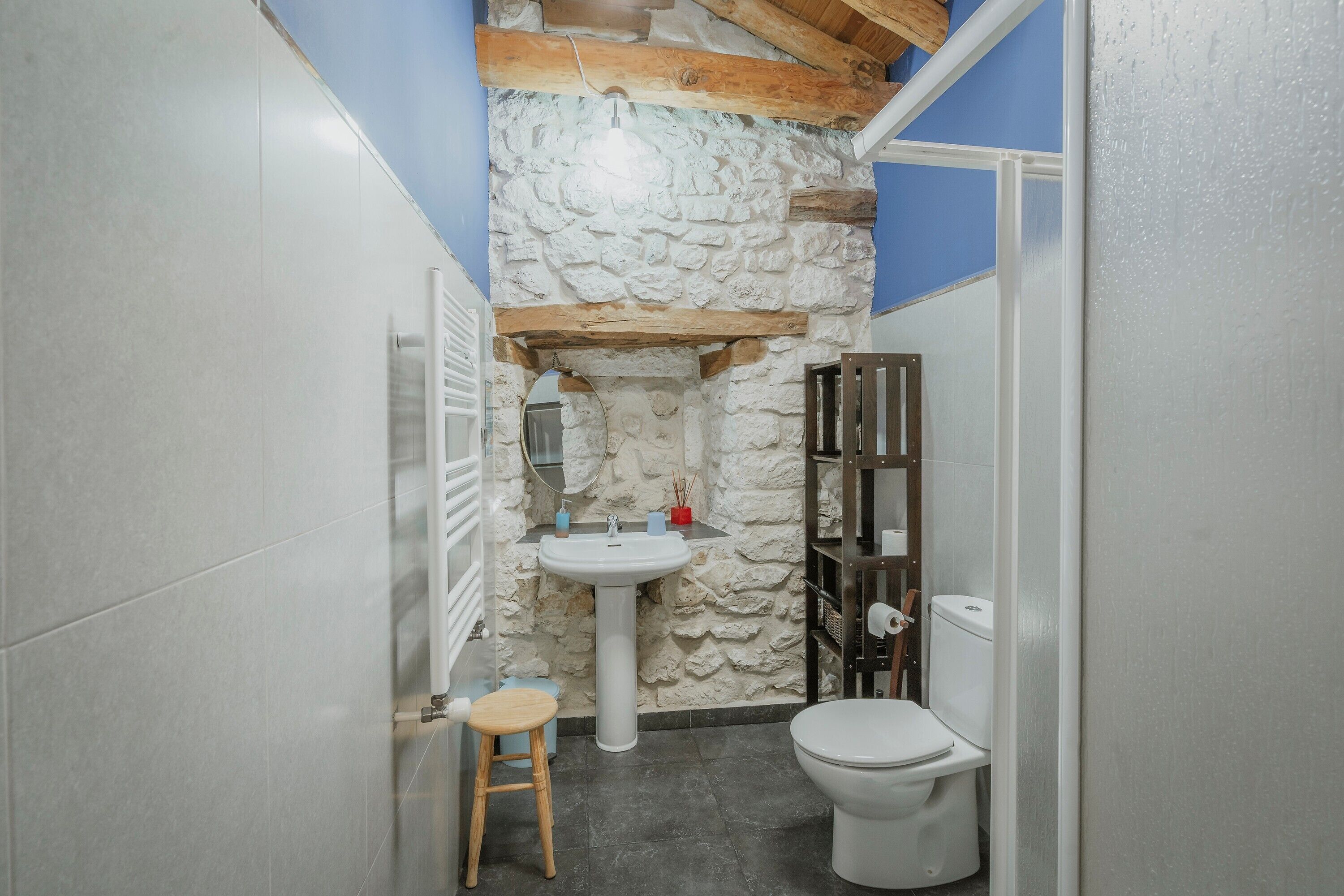 Ein Badezimmer mit Steinwand, einem Standwaschbecken, einer Toilette und einem hölzernen Hocker.