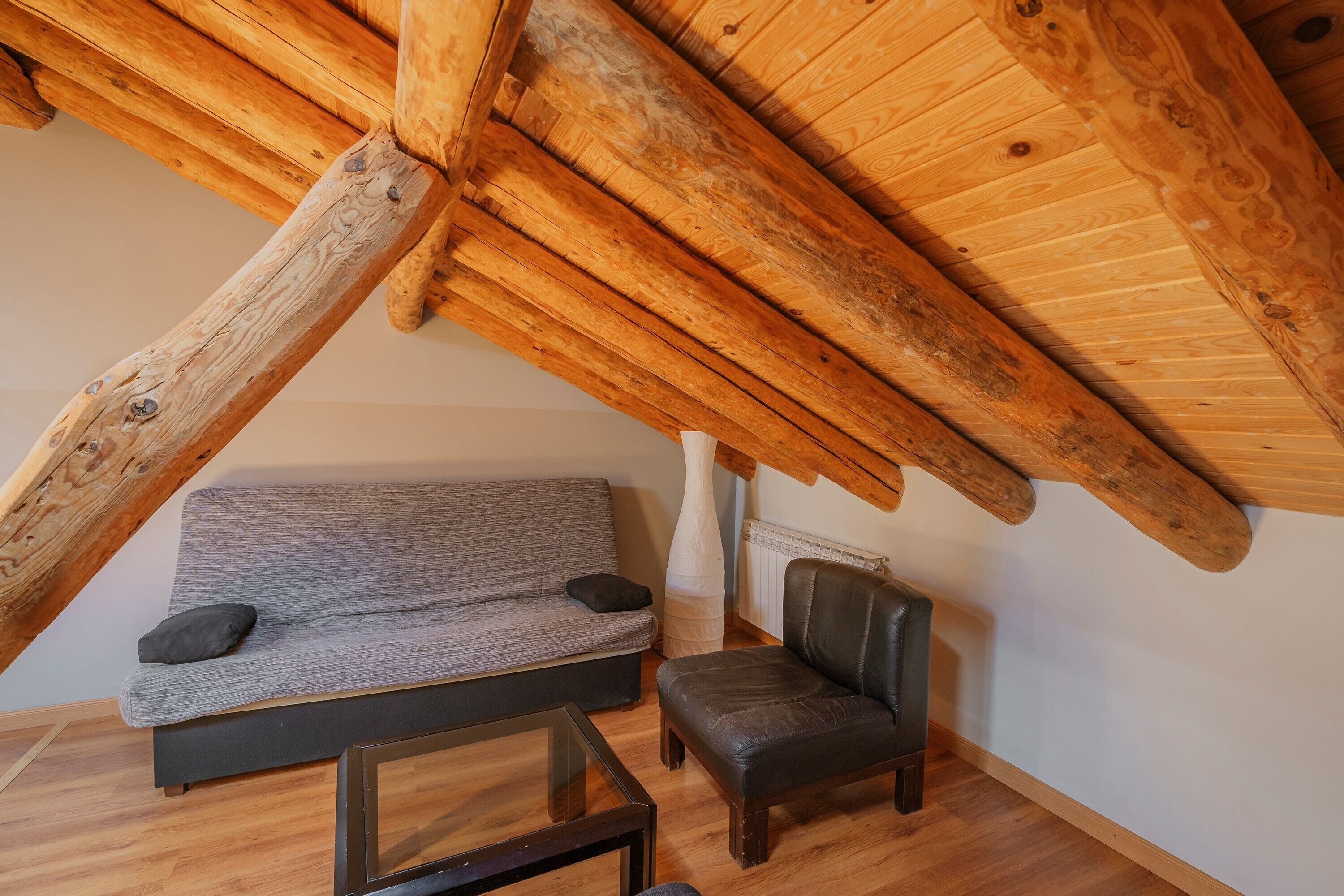 Una habitación estilo loft con vigas de madera a la vista, un sofá gris, un sillón de cuero y una mesa de centro de vidrio.