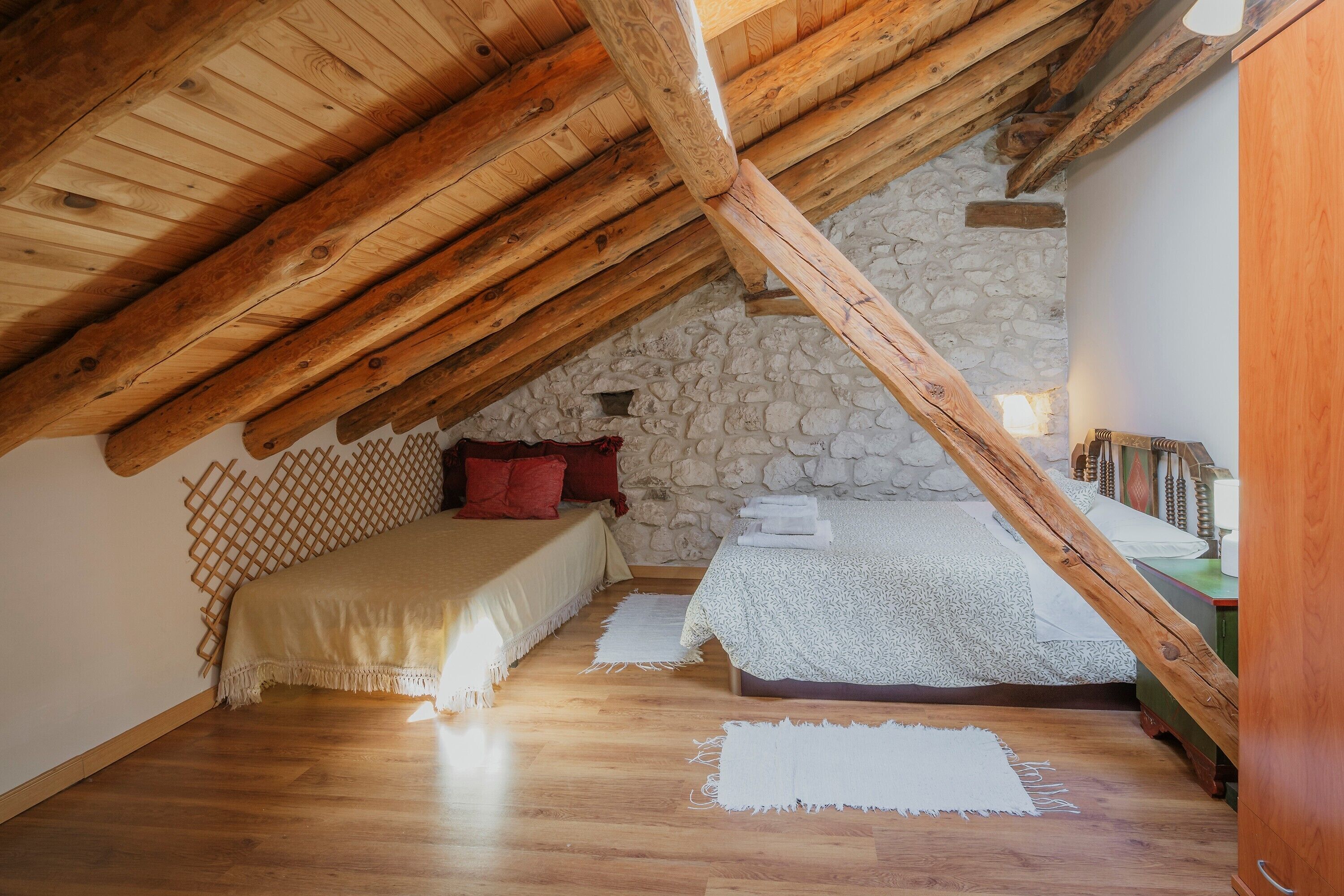 Un dormitorio con techo inclinado, vigas de madera, pared de piedra, dos camas y una pequeña mesa con lámpara.