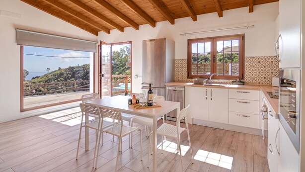A modern kitchen with wooden beams, a dining area with chairs, and a view through large windows. - Vacation Home 'Los Barranquillos' with Sea View, Private Pool, and Wi-Fi (Las Palmas)