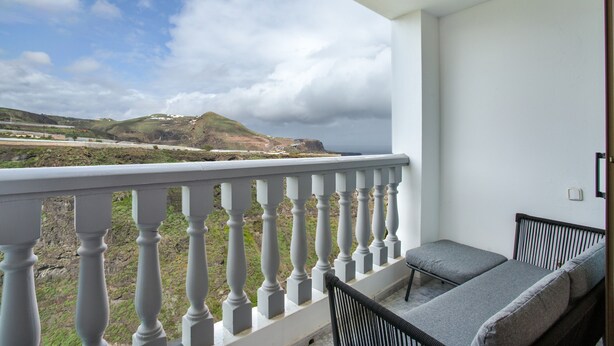 Un balcon doté d’une balustrade blanche, d’un canapé gris avec des coussins et offrant une vue sur un paysage côtier avec des collines et des nuages.