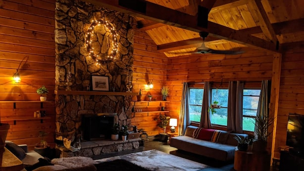 A cozy log cabin interior with a stone fireplace, wooden walls, a stone wall with a wreath, a sofa, and a dining area. - Rustic Modern Cabin w Hot Tub 1 5 hrs From NYC (Montgomery)