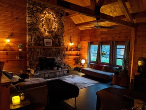 A cozy log cabin interior with a stone fireplace, wooden walls, a stone wall with a wreath, a sofa, and a dining area.