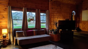 A cozy cabin interior with a wooden interior, a sofa with cushions, a television, and a window overlooking a green landscape. - Rustic Modern Cabin w Hot Tub 1 5 hrs From NYC (Montgomery)