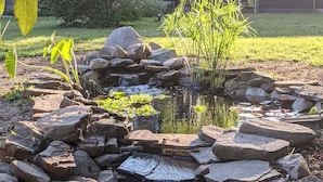 A pond with a waterfall, surrounded by rocks and plants, with a wooden cabin in the background. - Rustic Modern Cabin w Hot Tub 1 5 hrs From NYC (Montgomery)