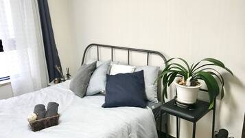 Ein ordentlich bezogenes Bett mit weißer Daunendecke, grau-blauen Kissen und einem kleinen Korb mit Hausschuhen. Auf einem Nachttisch neben dem Bett steht eine Topfpflanze.