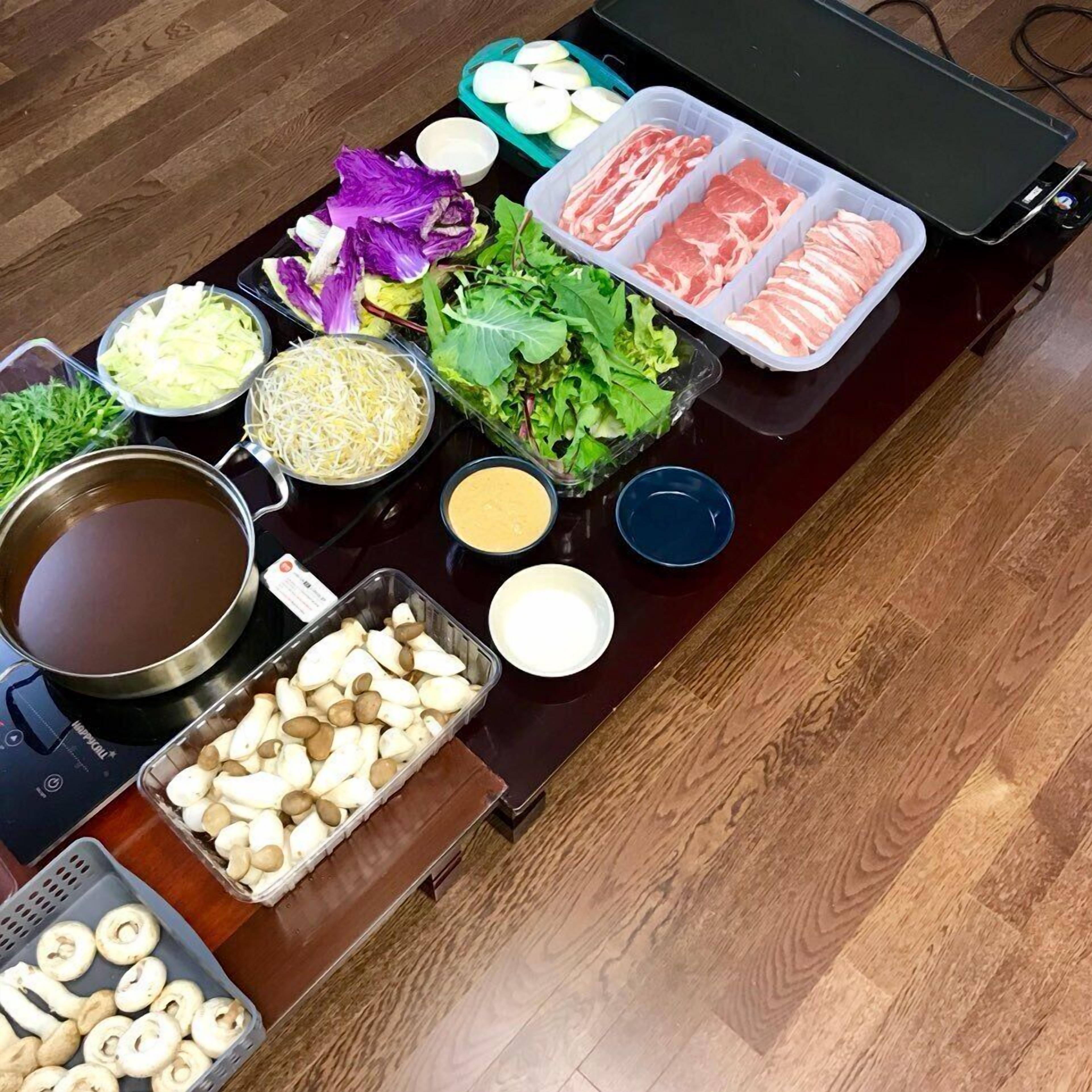 Een tafel met diverse etenswaren en toevoegingen, zoals plakjes vlees, groenten, champignons en een soepketel.