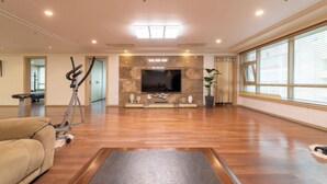 A spacious living room with a wooden floor, a large flat-screen TV mounted on a stone wall, a sofa, a treadmill, and a large window with blinds. - Sejong Party House  (Busan)