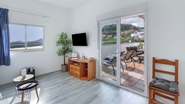 A room with a wooden desk, a television, a sliding glass door to an outdoor patio, and a view of a hilly landscape. - Los Adejo Vacation Home with Shared Heated Pool, Terrace, and Wi-Fi (Moya)