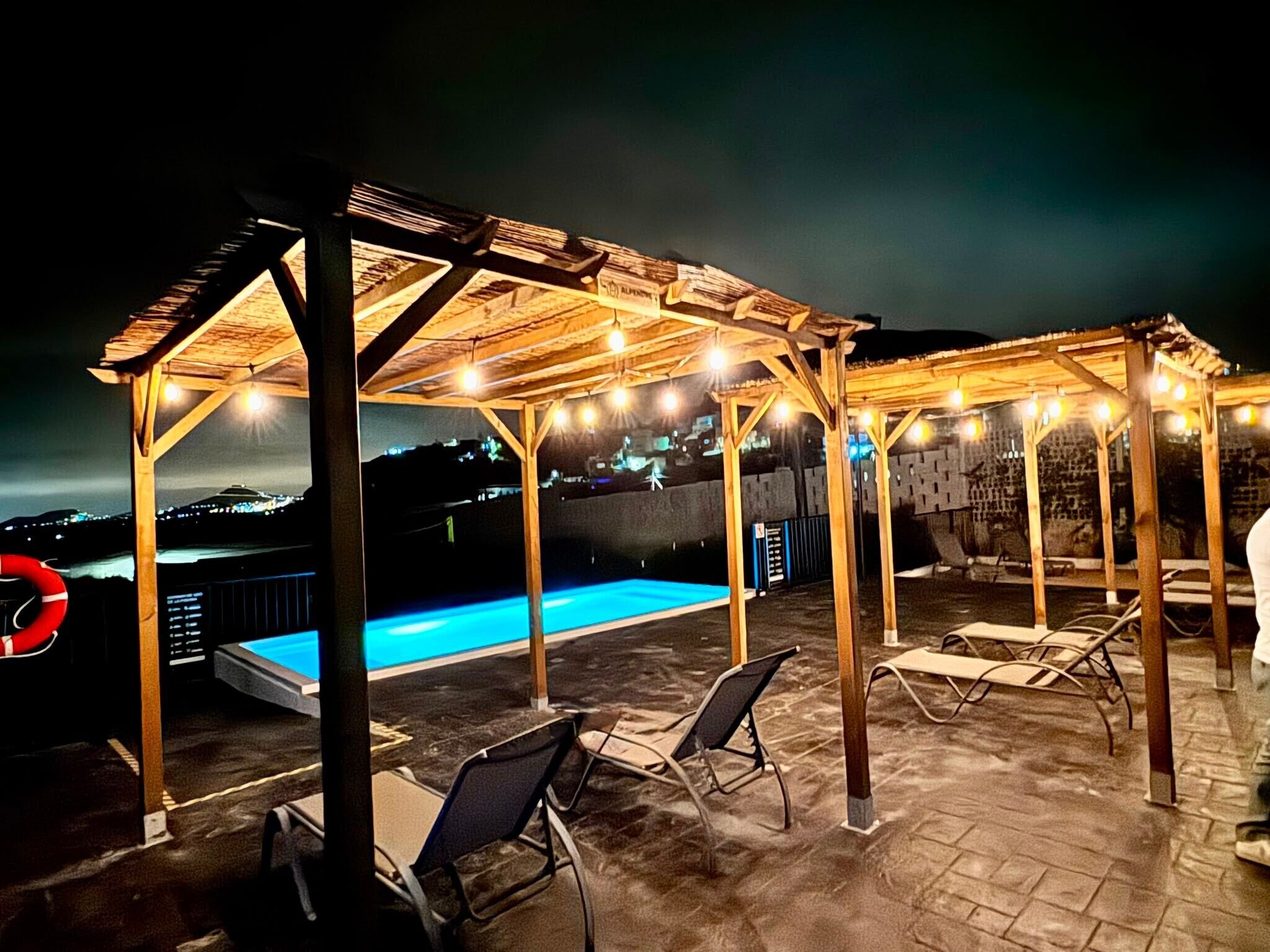 Een dakzwembad met houten pergola's, ligbedden en uitzicht op de stad 's nachts.