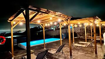 Een dakzwembad met houten pergola's, ligbedden en uitzicht op de stad 's nachts.