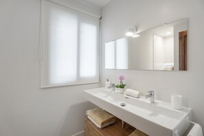 Un baño moderno con un espejo grande, un lavamanos blanco y una ventana con persianas.