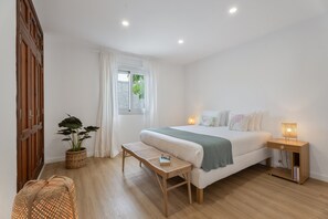 A bedroom with a bed, bedside table, bench, potted plant, and a window with curtains.